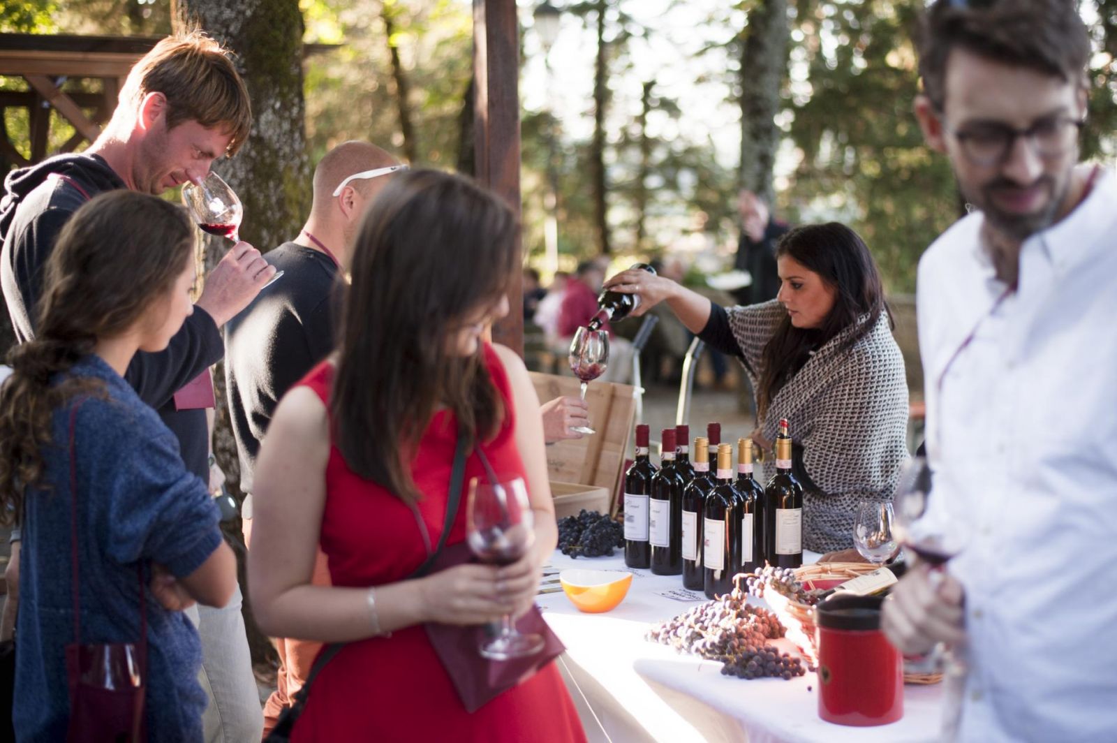 A busy scene at the Montefioralle Divino festival, showing people with wine glasses and a table with bottles and grapes