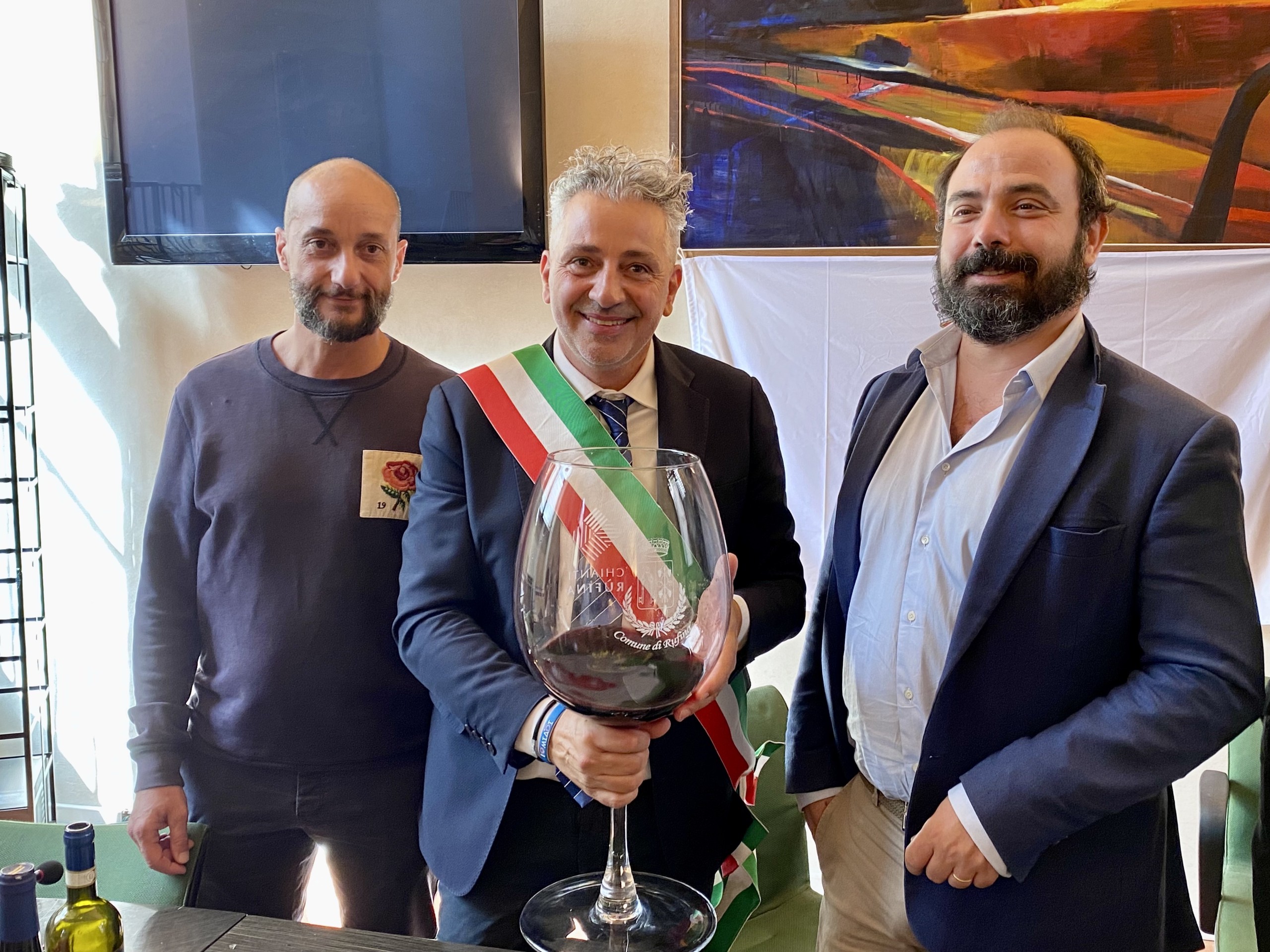 Three men posing for a photo, with the man in the center holding a giant glass of red wine and wearing an official Italian sash