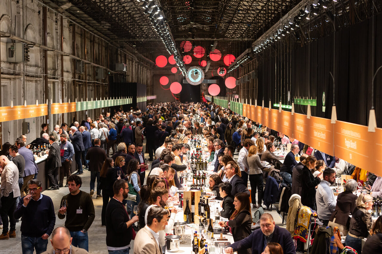 A large, long indoor hall filled with hundreds of people at long tables, tasting wine at the Expo del Chianti Classico