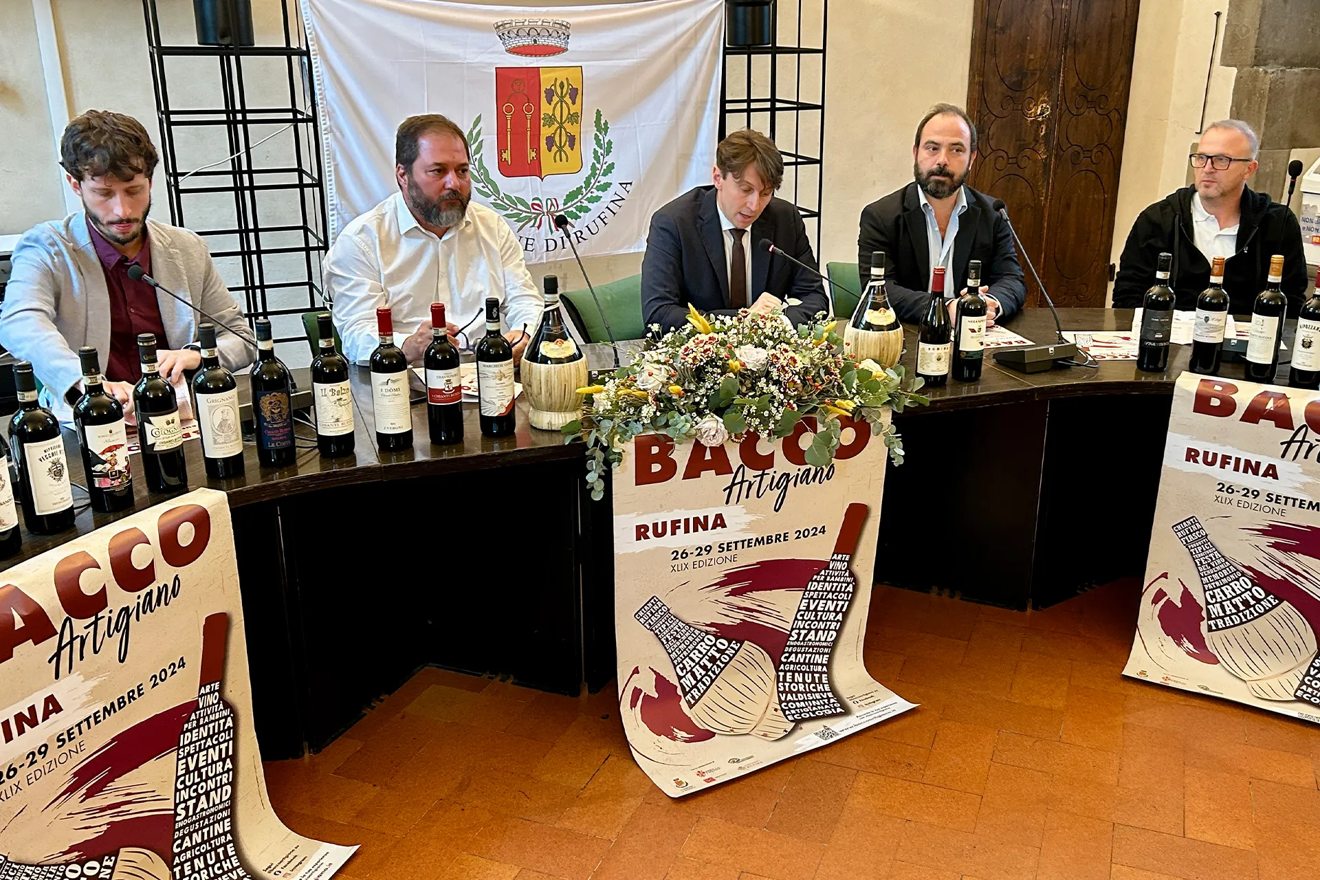 A panel of five men seated at a long table with a line of wine bottles and promotional posters for the Bacco Artigiano festival