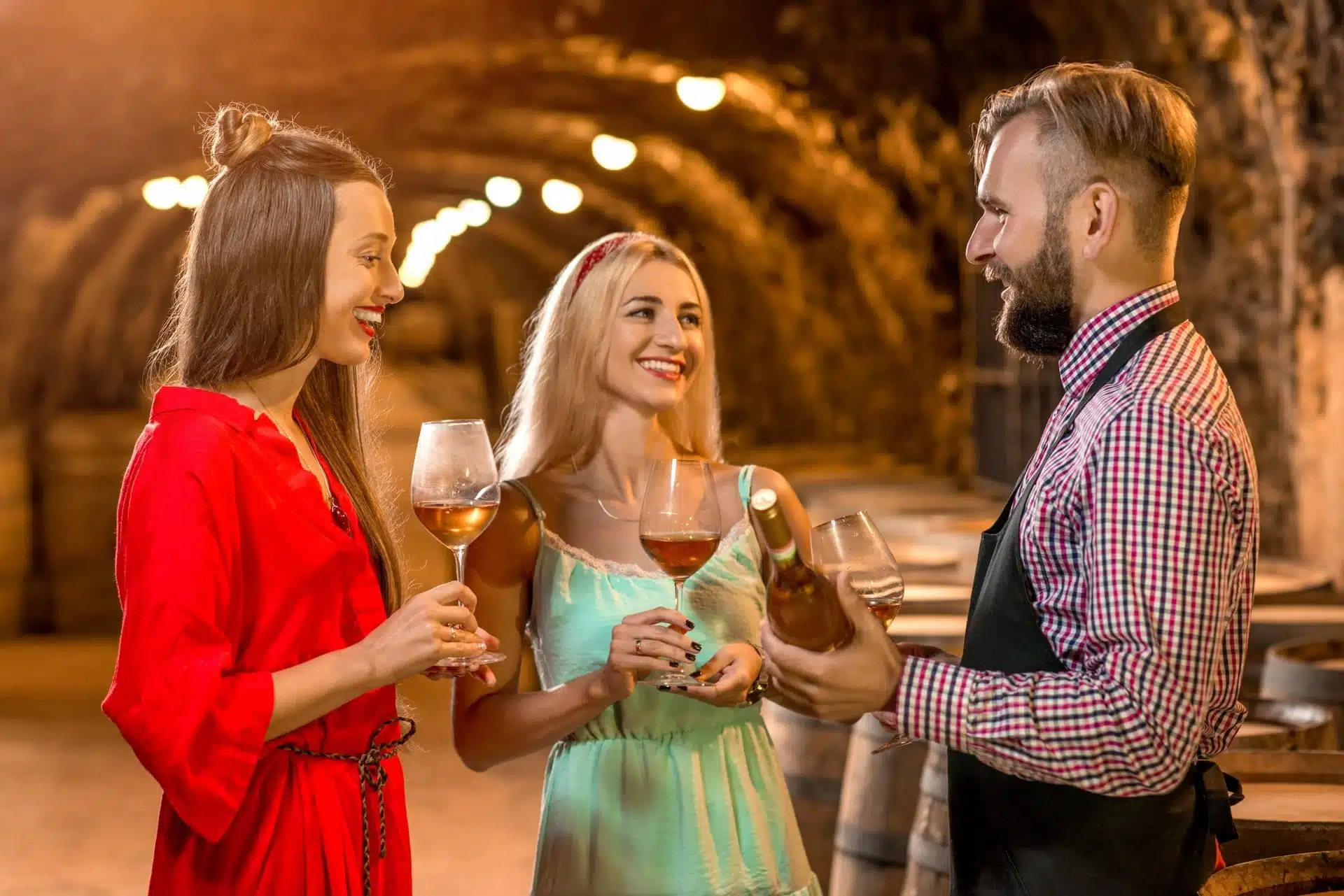 Two women tasting wine with a sommelier in a dimly lit wine cellar, featuring barrels and hanging lights