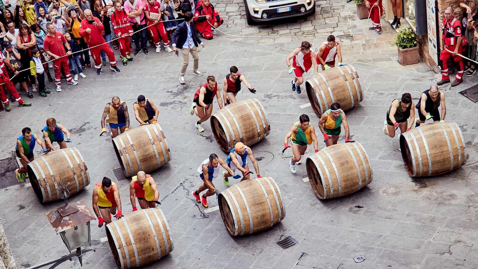 Men participating in a barrel-rolling race, with spectators lining the street and a car visible in the background