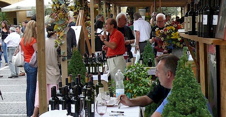 A busy scene at a wine-tasting event, showing attendees and vendors gathered around tables filled with bottles