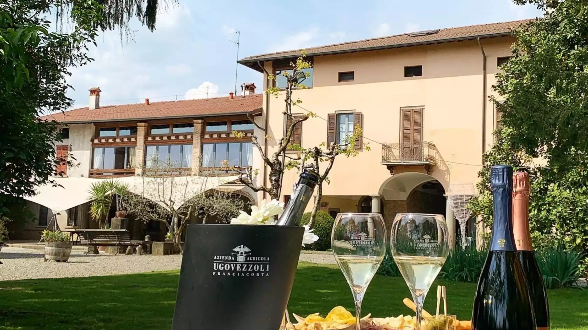 A pleasant outdoor scene at the Festa Delle Cantine, with a bottle of Franciacorta and snacks set up on a lawn in front of a winery