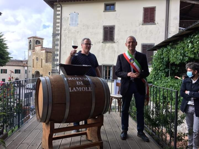 A man wearing an Italian sash standing with another man toasting with a glass of wine, in front of a wine barrel on a balcony