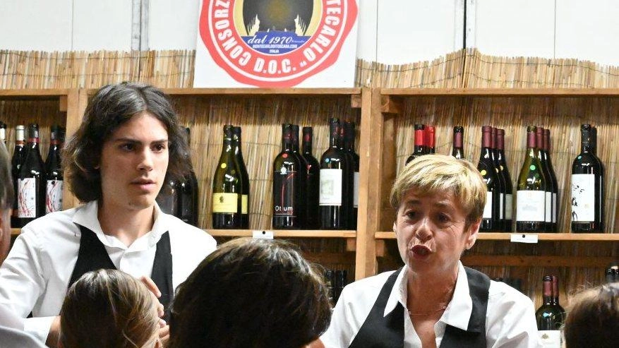 A man and a woman in vests speak to a crowd at a wine festival booth, with a selection of wine bottles on shelves behind them
