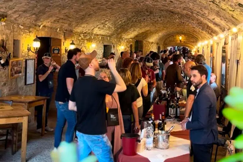 A long, crowded wine cellar with a vaulted stone ceiling, filled with people tasting wine at a long table