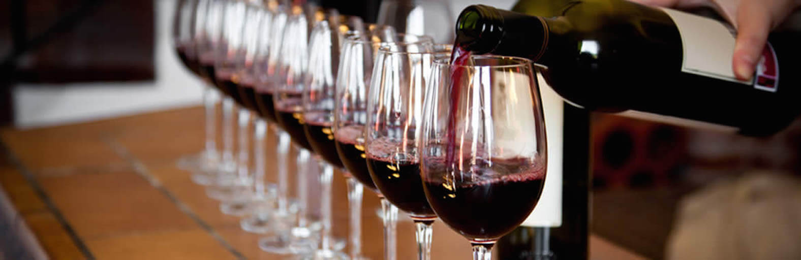 A person's hand pouring red wine from a bottle into a row of wine glasses at a tasting event