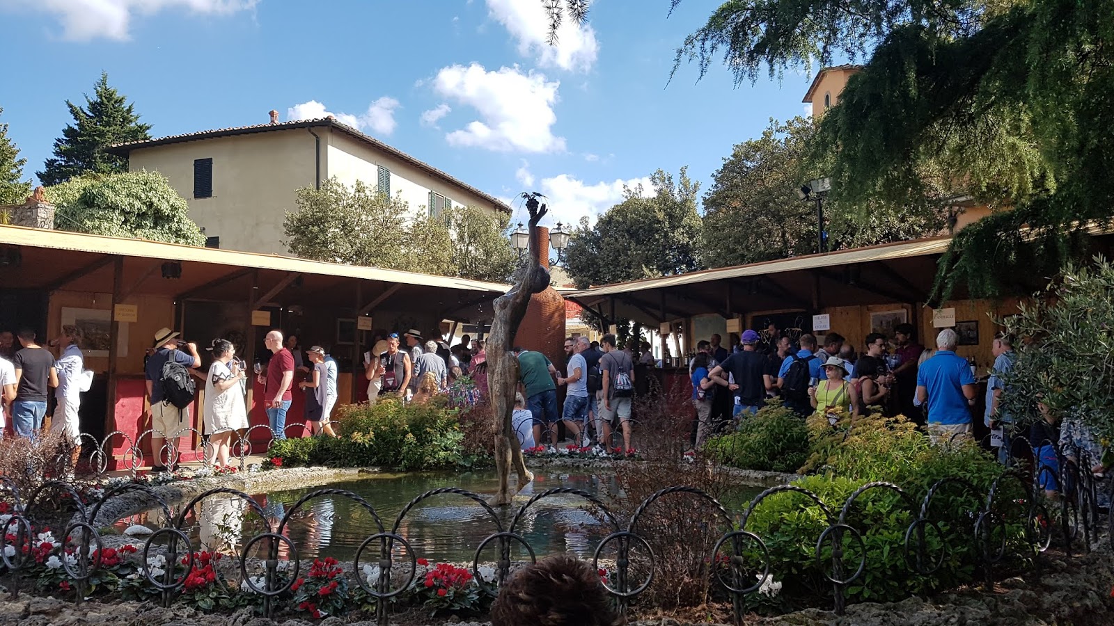 Guests socializing and tasting wine at an event with a decorative fountain in the center and buildings in the background